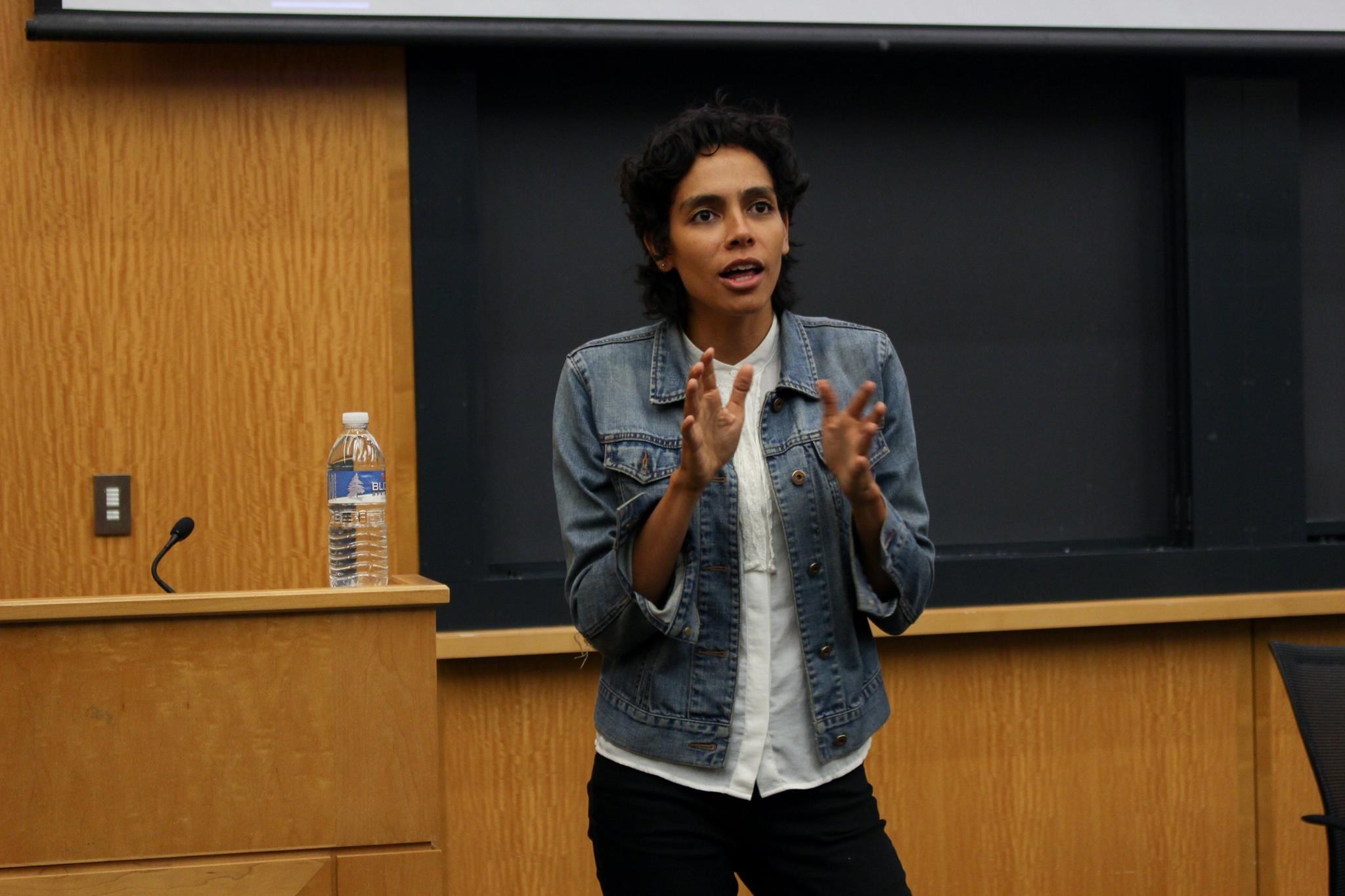 Frau am Rednerpult mit Mikrofon und Flasche, die vor einem Projektionsschirm eine Zuschauergruppe anspricht, mit einem Stuhl rechts und einer hölzernen Wand im Hintergrund.