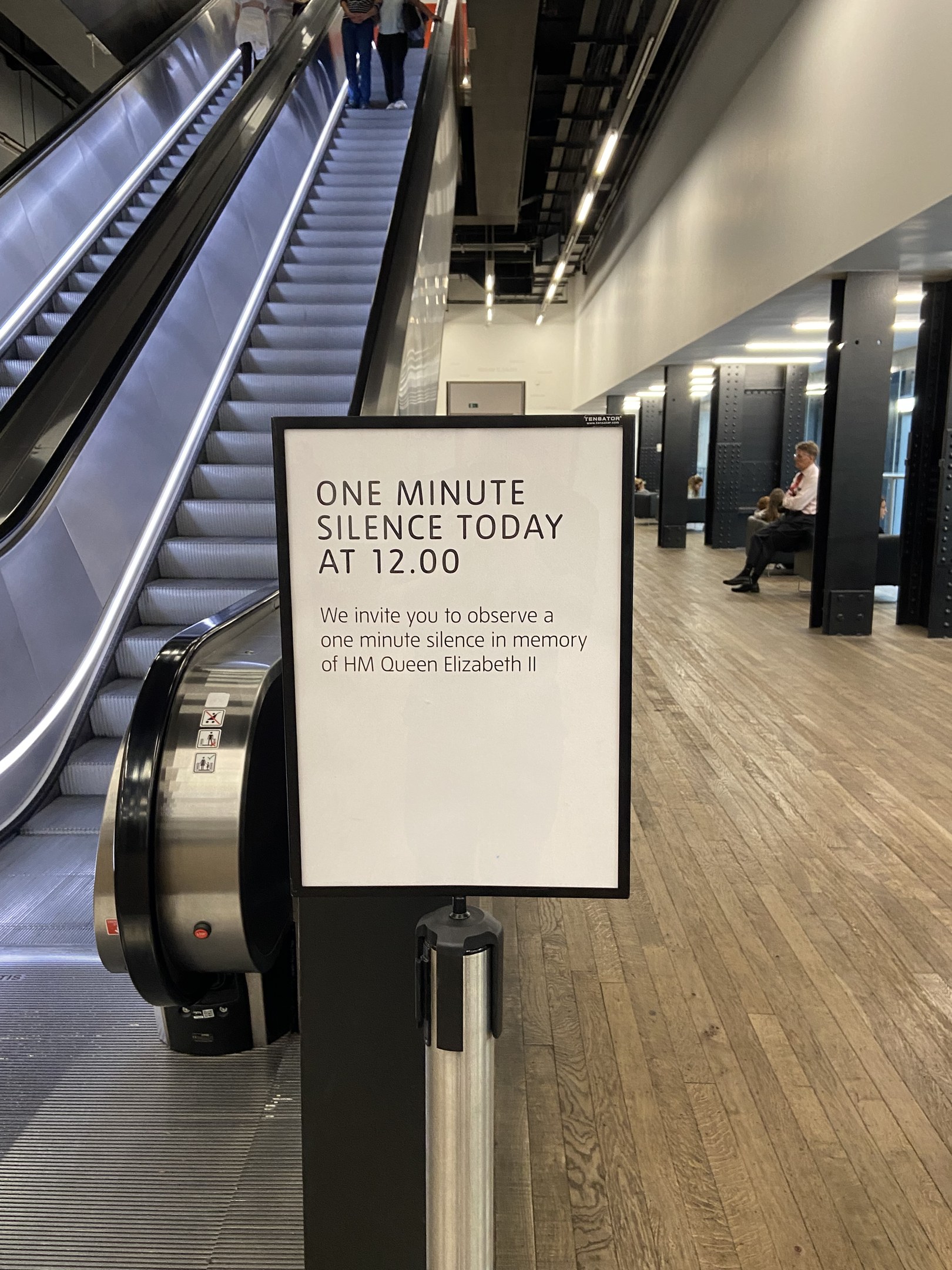 Eine Rolltreppe in einem Flughafen mit einem Schild, auf dem "Eine Minute Stille heute" steht, einige Menschen darauf und an der Decke angebrachte Lichter im Hintergrund.