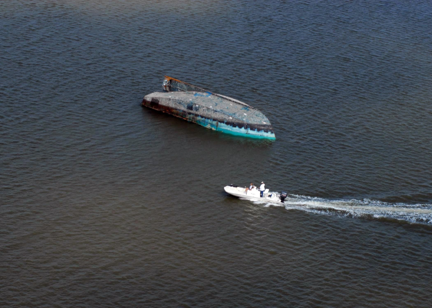 Ein kleines, teilweise untergetauchtes Wrackboot, das einen Fluss hinunterfährt, neben einem größeren Boot mit ein paar Menschen an Bord.