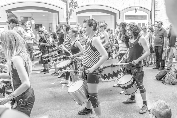Eine Gruppe von Menschen, die draußen Musikinstrumente spielen, vor einer geschlossenen Glastür mit einem Licht im Hintergrund.