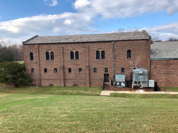 Ein großes Backsteingebäude mit einem grünen Rasen, Bäumen und einem klaren blauen Himmel, mit Fenstern, einer Tür und einer Treppe, die hinaufführt, und einer Restaurierungsmaschine auf der rechten Seite.