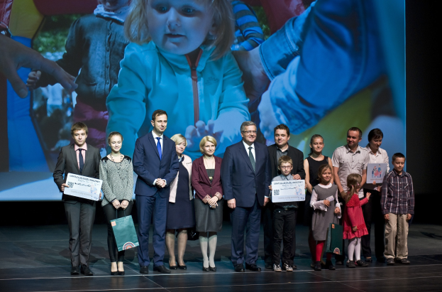 Gruppe von Menschen auf einer Bühne mit Taschen und Urkunden, mit einem Bildschirm, der ein Bild eines Kindes und andere Personen im Hintergrund anzeigt.