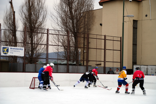 Menschen beim Eishockeyspielen auf einem Eis mit Gebäuden, Bäumen, einer Straßenlaterne, einem Namensschild und Zäunen im Hintergrund bei klarem Himmel.