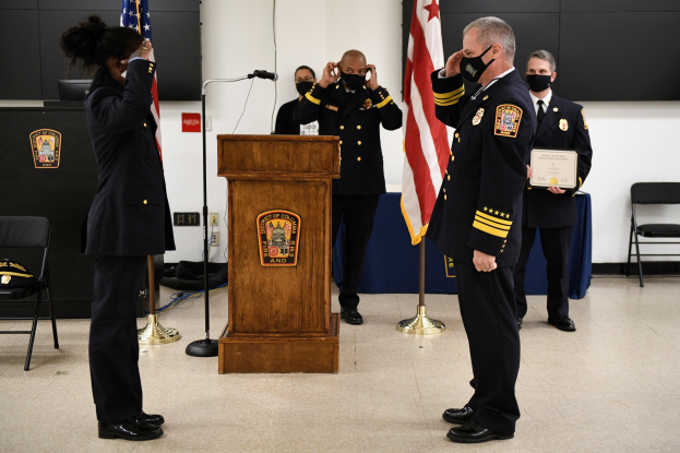 Gruppe uniformierter Personen in Masken auf einem Podium mit einem Mikrofon, umgeben von Flaggen und Möbeln, während einer Vereidungszeremonie für einen neuen Feuerwehrchef.