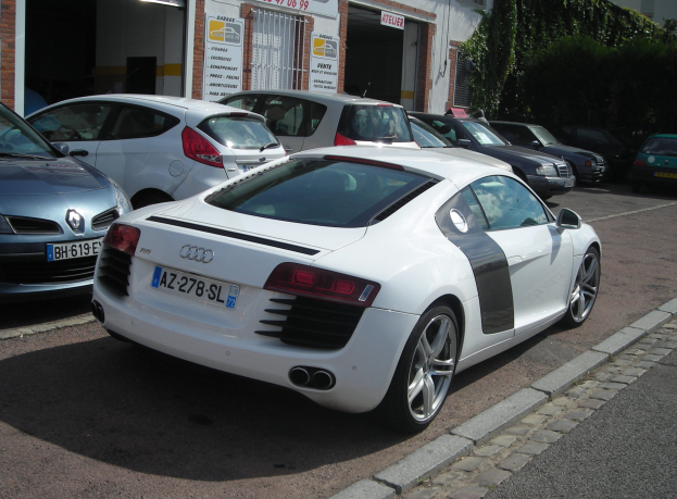 Ein weißer Audi R8, der auf einer Straße vor einem Gebäude mit Texttafeln geparkt ist, umgeben von Bäumen auf der rechten Seite.