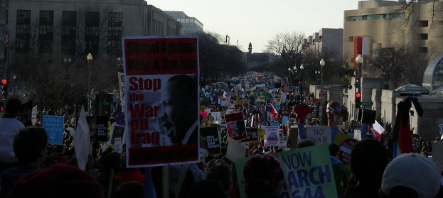 Menschen mit Plakaten auf einer Straße, mit Gebäuden, Bäumen, Laternenmasten und dem Himmel im Hintergrund.