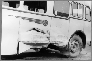 Schwarzes und weißes Foto eines beschädigten Busses am Straßenrand mit eingedellter Front, zersplitterten Fenstern und einer offenen Fahrertür, die das Innere zeigt.