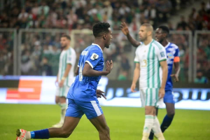 Ein Fussballspieler in einem blauen und weissen Trikot feiert nach einem Tor, umgeben von einer Gruppe von Menschen in weissen und grünen Trikots, mit einer weissen und blauen Tafel und einem Geländer im unscharfen Hintergrund.