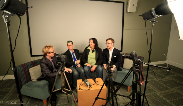 Eine Gruppe von drei Personen sitzt auf einer Couch und gibt ein Interview. Links sitzt eine Person in einem schwarzen Mantel und Hose, in der Mitte eine Person in einem Mantel und blauer Krawatte mit einem Mikrofon und rechts eine strahlende Person. Hinter ihnen ist ein Projektionsschirm, auf dem Tisch in der Mitte ein Mikrofonrecorder und auf beiden Seiten Kameras mit großen Scheinwerfern.