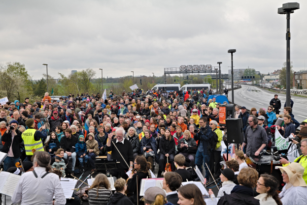 Große Menschenmenge marschiert vor einer Menge mit Musikinstrumenten, Ständern und anderen Gegenständen, mit Fahrzeugen, Laternenmasten, Bäumen, Gebäuden und einem bewölkten Himmel im Hintergrund, wahrscheinlich während einer Demonstration in Manchester.