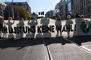 Eine Gruppe von Menschen mit Masken hält ein Transparent mit der Aufschrift "Wir sind die letzten keine wahl" vor Gebäuden, Bäumen, Laternen, Ampeln und einem Zelt, mit dem Himmel im Hintergrund.