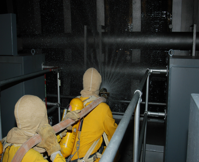 Zwei Feuerwehrleute in gelben Uniformen, Helmen und Handschuhen spritzen Wasser aus einem Schlauch auf ein Metallgeländer, mit Metallstangen und anderen Gegenständen im Hintergrund und Text unten.