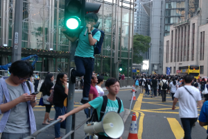 Ein Mann steht auf einer Ampel und hölt ein Megafon und ein Handy, umgeben von einer Gruppe von Menschen auf der Straße, mit Verkehrskegeln, einem Metallzaun, einer Hinweistafel, Fahrzeugen, Gebäuden, Bäumen und einem bewölktem Himmel im Hintergrund.