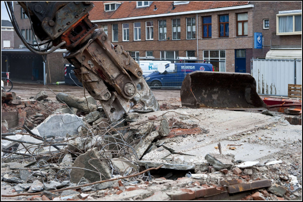 Bagger bei einem Schutthaufen mit verstreuten Trümmern, Fahrzeugen auf der Straße, Gebäuden im Hintergrund und Text unten.