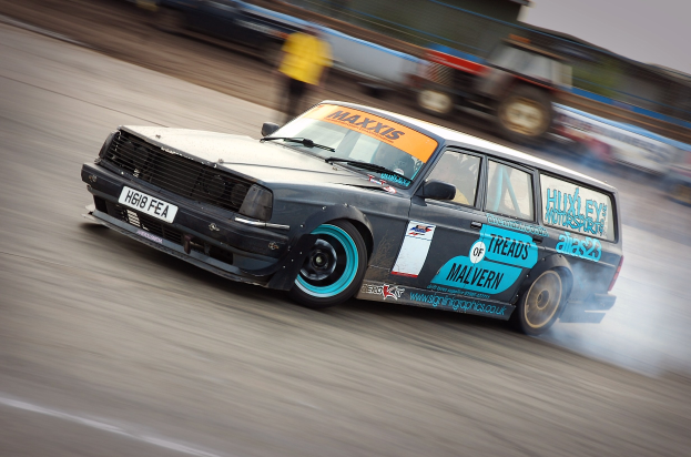 Ein Rennwagen fährt mit hoher Geschwindigkeit auf einer Rennstrecke, aus dem Rauch austritt, mit anderen Fahrzeugen und Zuschauern im unscharfen Hintergrund.