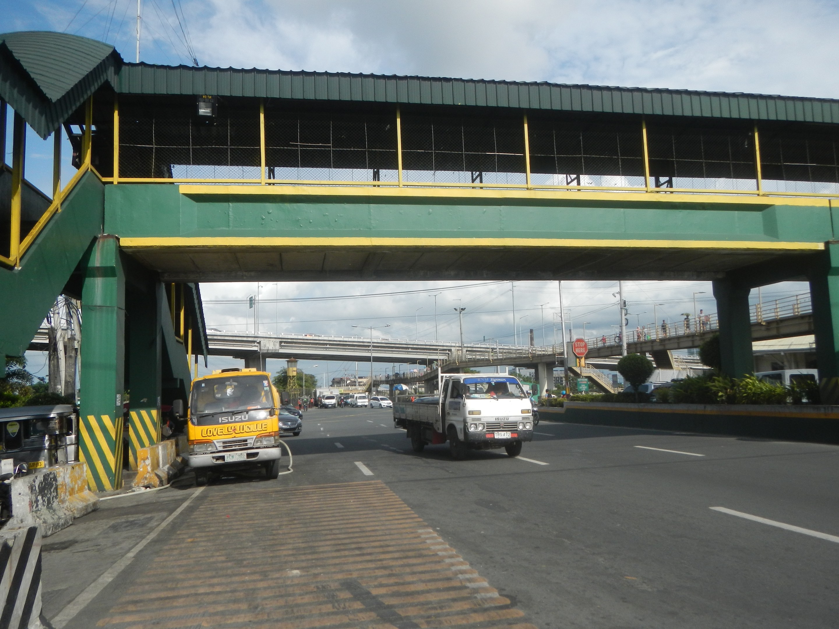 Mautstelle mit Fahrzeugen auf einer Straße unten, eine Brücke mit Geländern und Säulen, Laternenmäste, Schilder, Bäume und ein bewölkter Himmel im Hintergrund, mit einem Lastwagen, der neben der Brücke fährt.