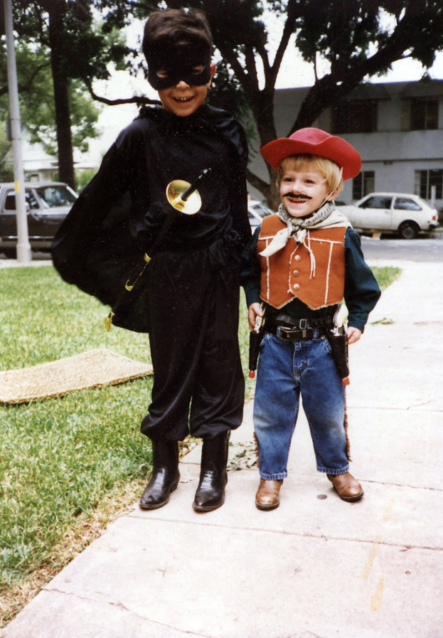 Kinder in verschiedenen Kost√ľmen auf einer Stra√e mit Fahrzeugen, Geb√§uden, B√§umen und Stra√enmasten im Hintergrund unter einem Himmel.