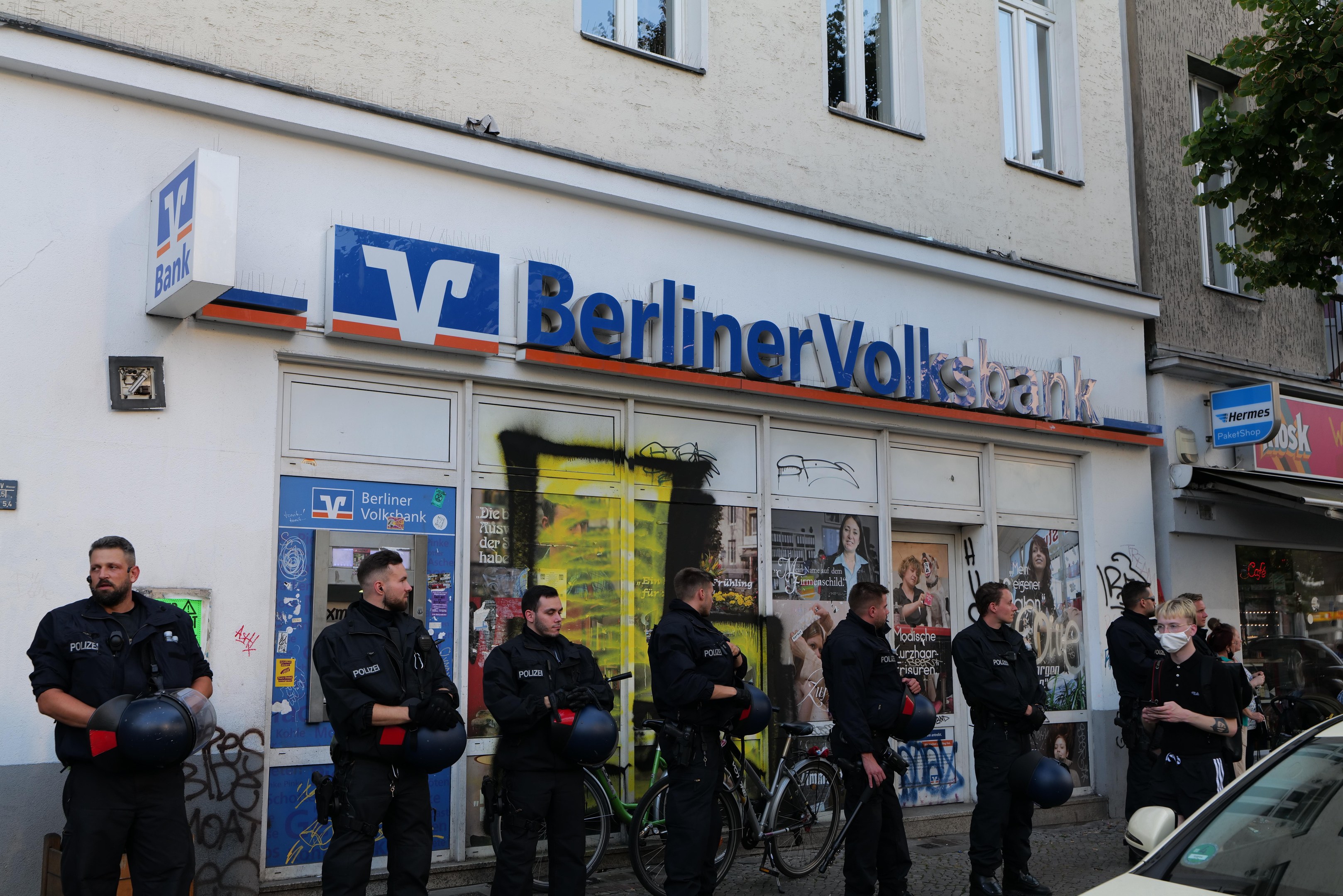 Polizisten in Uniform vor einer Berliner Volksbank-Filiale mit Helmen in den Händen stehend; rechts ein geparktes Auto und links ein Baum; im Hintergrund Gebäude mit Fenstern und Texttafeln