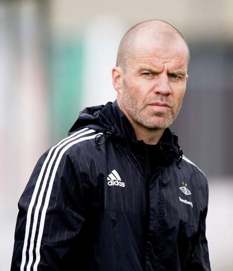 Baldiger Trainer in marineblauer Jacke mit weißen Streifen und weißem Kragen auf einem Fußballfeld mit verschränkten Händen und ernster Miene.