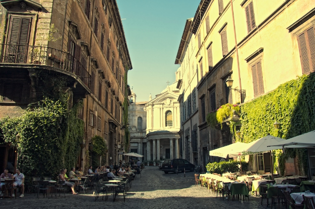 Ein Weg mit Tischen, Stühlen und parkenden Autos, gesäumt von Gebäuden mit Pflanzen im Vordergrund und Himmel darüber.