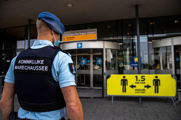 Ein Polizist in blauer Uniform und Maske steht vor einem gläsernen Gebäude, mit einem gelben Schild rechts daneben und zusätzlichen Schildern im Hintergrund.