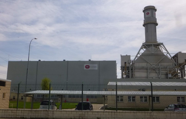 Ein großes Gebäude mit einem zentralen Turm, umgeben von einem Zaun, mit Fahrzeugen auf der benachbarten Straße, Polen, Lampen, Drähten, Bäumen und Gras im Vordergrund und einem bewölkten Himmel im Hintergrund, das eine neue Energiespeicheranlage darstellt.