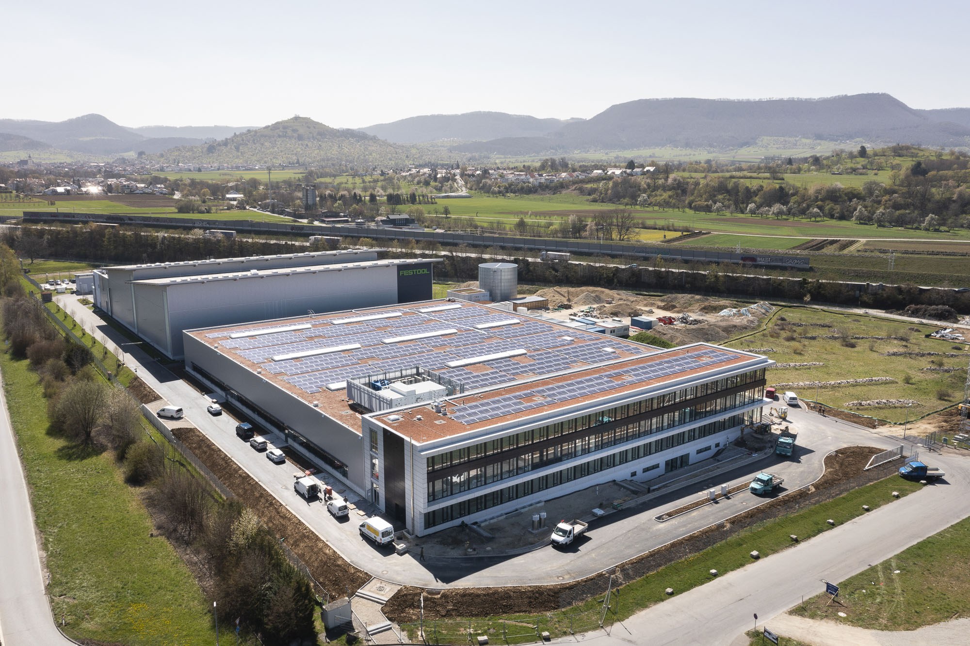 Luftaufnahme eines großen Fabrikgebäudes mit Solarmodulen auf dem Dach, umgeben von Fahrzeugen, Bäumen, Gras und Masten, mit Hügeln und einem klaren blauen Himmel im Hintergrund.