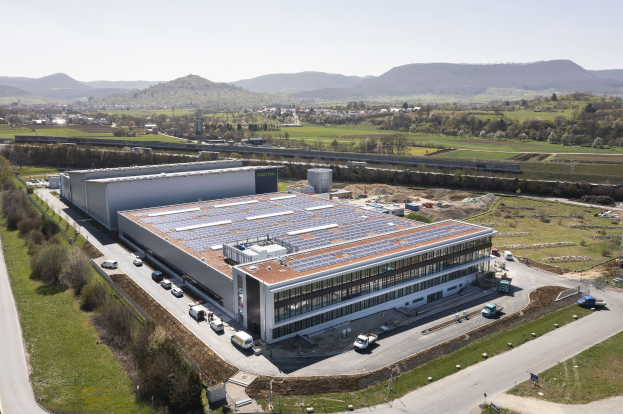 Luftaufnahme eines großen Fabrikgebäudes mit Solarmodulen auf dem Dach, umgeben von Fahrzeugen, Bäumen, Gras und Masten, mit Hügeln und einem klaren blauen Himmel im Hintergrund.