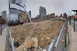 Baustelle mit einem großen Schutthaufen, umgeben von Gebäuden, Straßeninfrastruktur, Fahrzeugen, Fußgängern, Bäumen und einem klaren Himmel.