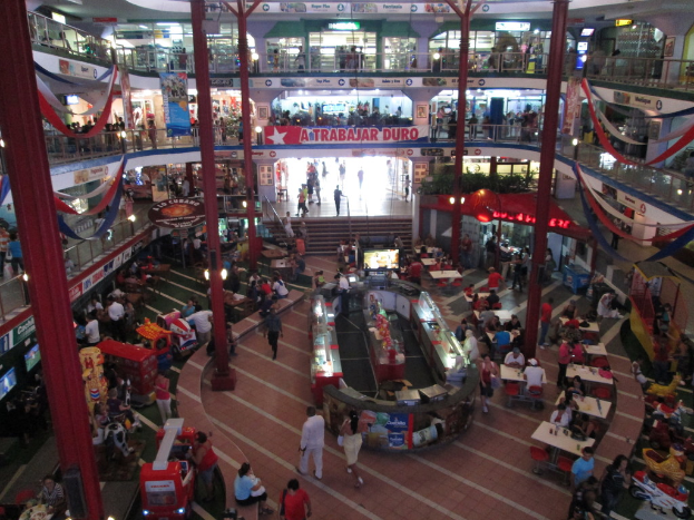 Ein belebtes Einkaufszentrum mit Säulen, Menschen, Tischen, Fliesen, Flaggen, Geschäften, einer Fahne, Treppen und einem Namensschild.