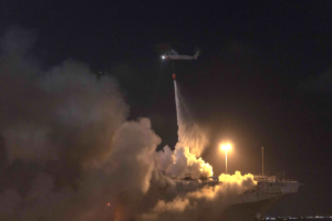 Ein Hubschrauber fliegt über ein großes Schiff mit rauchender Luke, beleuchtet von den Lichtern des Schiffs und dem Himmel im Hintergrund.