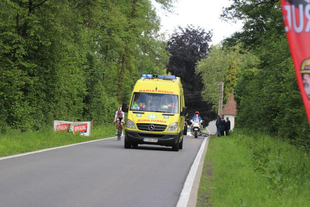 Ambulanz fährt auf einer Straße mit Fahrradfahrern daneben, Gras und Bäume auf beiden Seiten, Häuser und Strommasten im Hintergrund unter einem klaren blauen Himmel.