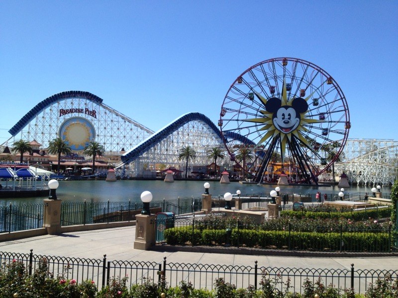 Disneylands California Adventure Vergnügungspark mit einem Riesenrad, Achterbahn, Grünflächen, Wasser, Laternenmasten und einem hellblauen Himmel.