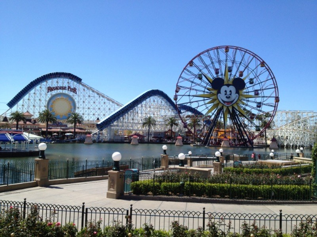 Disneylands California Adventure Vergnügungspark mit einem Riesenrad, Achterbahn, Grünflächen, Wasser, Laternenmasten und einem hellblauen Himmel.