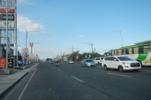 Eine belebte Stadtstraße voller Autos, Busse, Motorräder und Gebäude, mit einem Gehweg auf der linken Seite und verschiedenen städtischen Elementen im Hintergrund.