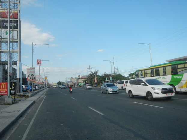 Eine belebte Stadtstraße voller Autos, Busse, Motorräder und Gebäude, mit einem Gehweg auf der linken Seite und verschiedenen städtischen Elementen im Hintergrund.