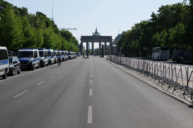 Lange Reihe von Polizeiwagen entlang einer Straße vor dem Brandenburger Tor mit Menschen auf Fahrrädern und Barrieren, Bäume säumen die Seiten und ein Bogen mit Statuen im Hintergrund.