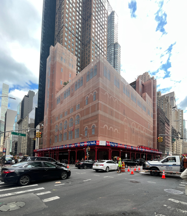 Eine belebte Stadtstraße mit hohen Gebäuden, Fahrzeugen, Fußgängern und diverser Straßenmöblierung, mit dem Hall in New York City prominent in der Mitte, unter einem bewölkten Himmel mit einem Wasserzeichen.