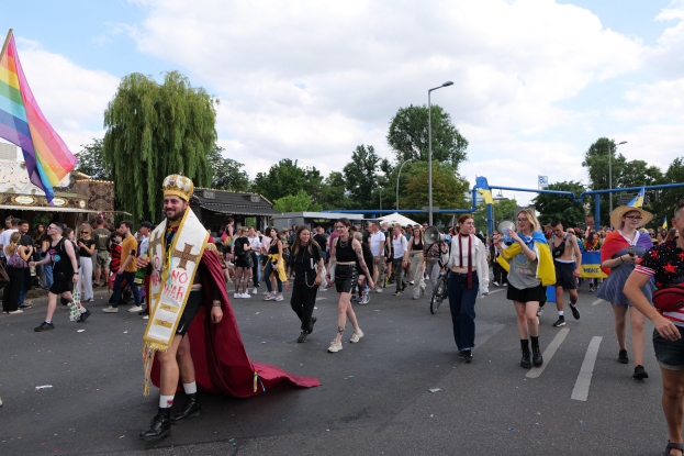 Eine Gruppe von Menschen marschiert bei der Christopher Street Day Parade 2018, einige mit Musikinstrumenten und andere mit Mützen, die eine Regenbogenflagge tragen, während sie eine Straße mit Laternenpfählen, Bäumen, Schuppen und einem bewölkten Himmel entlanggehen.