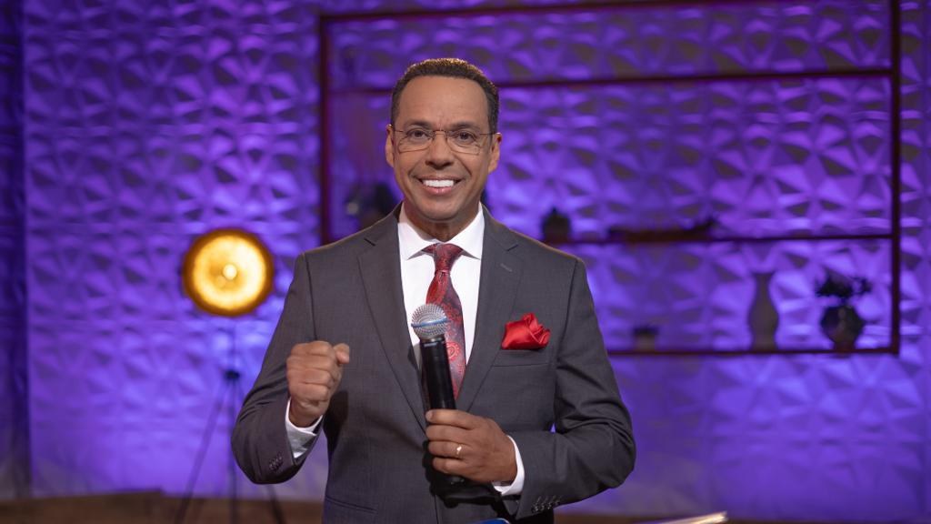 Stephen Colbert, Moderator von *The Daily Show with Trevor Noah*, steht auf einer Bühne und hält ein Mikrofon, trägt einen Anzug, Krawatte und Brille, hinter ihm ist ein Scheinwerfer und Ständer zu sehen.