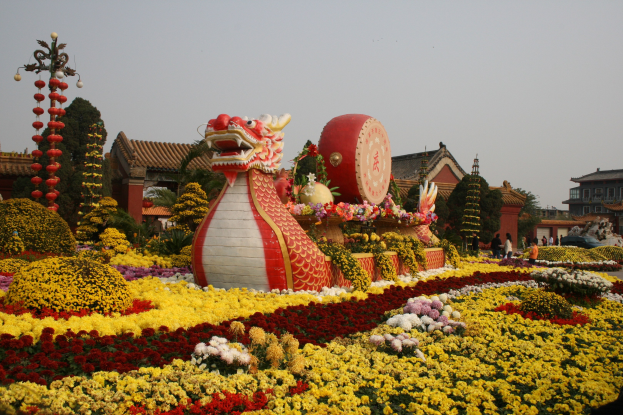 Ein farbenfrohes chinesisches Neujahrsparade mit einer Drachenstatue inmitten von bunten Blumen in Gelb, Rot, Pink und Weiß, umgeben von Gebäuden, Bäumen und einem klaren blauen Himmel, und Menschen, die umhergehen.