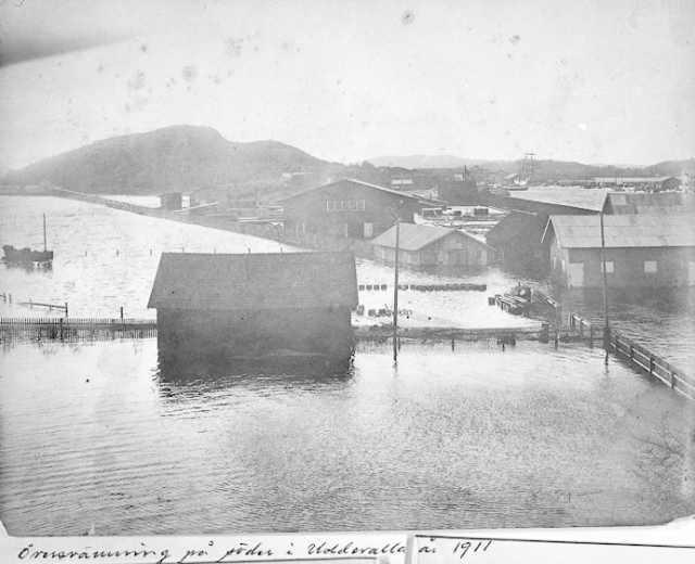 Schwarz-weiß-Foto einer kleinen Stadt mit Häusern, Strommasten und teilweise in Überschwemmungswasser versunkenen Booten, umgeben von Hügeln unter einem klaren Himmel, beschriftet mit "1911 Überschwemmungsschaden in St. Petersburg."
