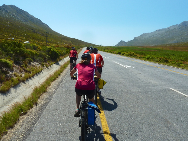Eine Gruppe von Menschen in Helmen auf einer Straße radelnd, mit Pflanzen, Gras, Polen, Bergen und einem bewölkten Himmel im Hintergrund.