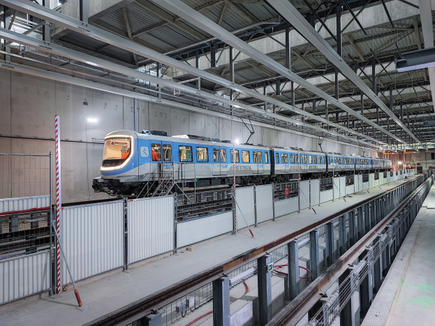 Blauer und weißer Zug fährt durch eine große Bahnhofshalle mit einem Bahnsteig auf der rechten Seite, einer Wand im Hintergrund und einer Decke mit Stangen und Lampen.