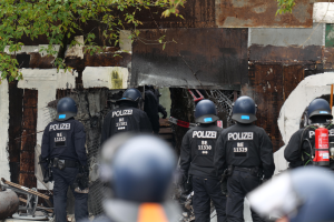 Eine Gruppe von Polizisten in schwarzen Uniformen und Helmen steht vor einem Gebäude, mit einem Feuerlöscher auf der rechten Seite und Bäumen im Hintergrund.