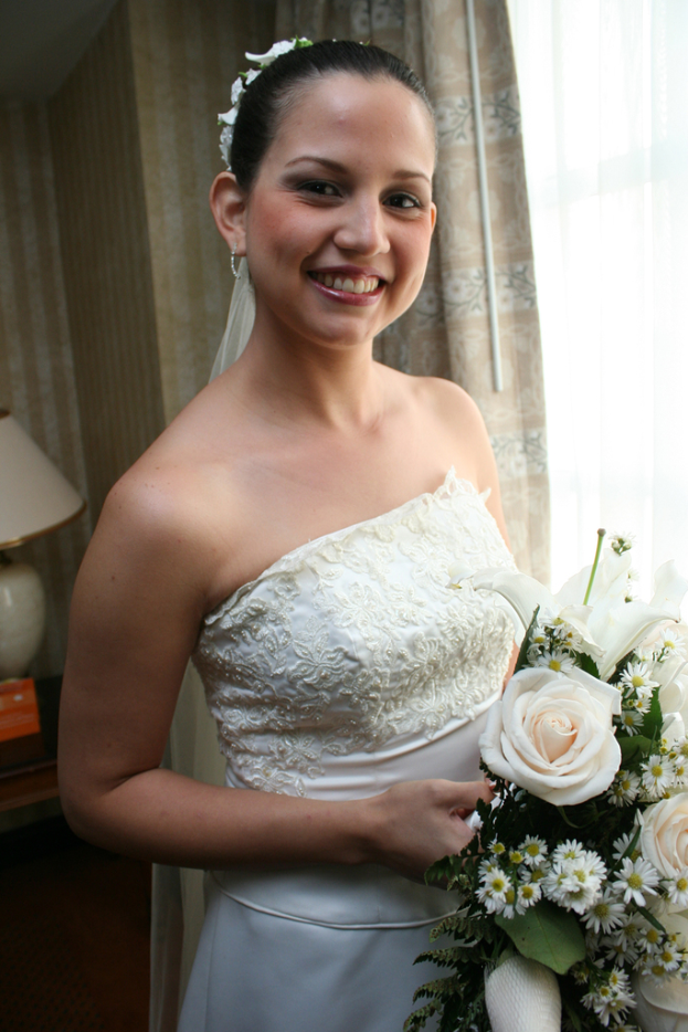 Eine strahlende Braut steht in der Mitte, hält einen Blumenstrauß und hat einen Vorhang und eine Wand im Hintergrund sowie eine Lampe auf einem Tisch links.