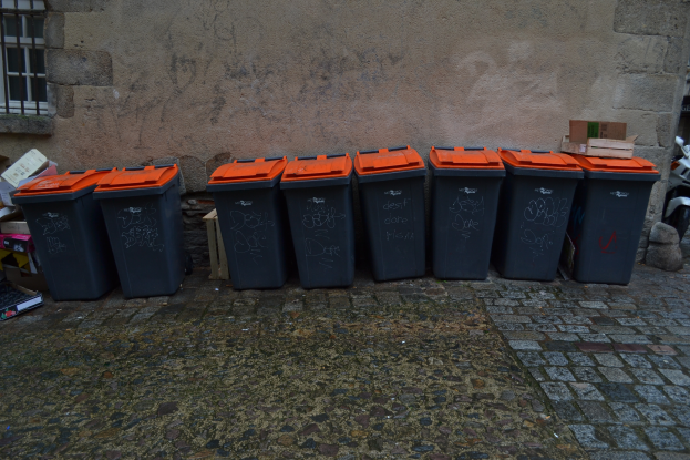 Eine Reihe schwarzer und oranger Mülltonnen auf einem Kopfsteinpflasterweg, mit verstreuten Kartonagen und anderen Gegenständen links, einer Wand mit einem Fenster im Hintergrund und einem Roller auf der rechten Seite.