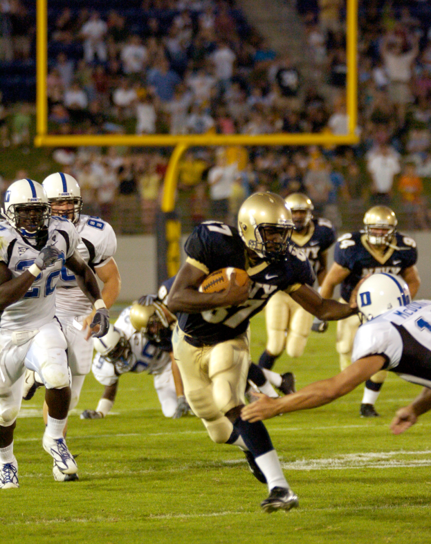 Ein Füßballspieler in einem Sportsdress und Helm läuft mit dem Ball, umgeben von anderen Spielern, mit unscharfen Hintergrundelementen und Text unten.