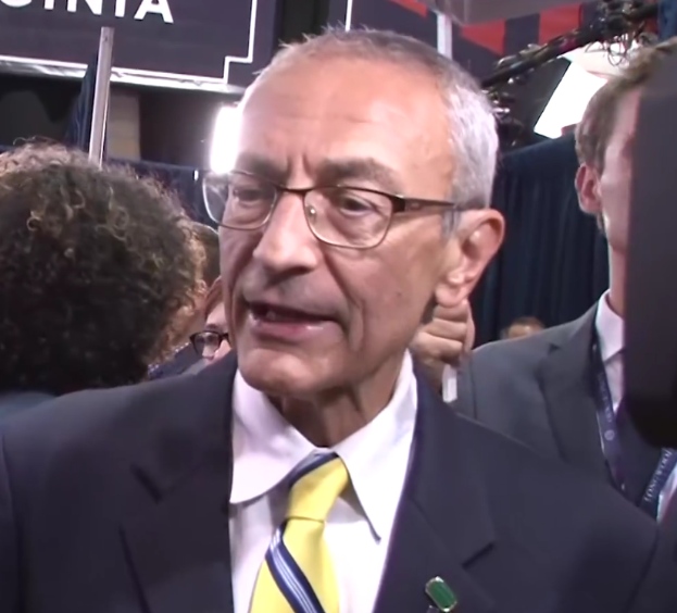 Ein Mann im Anzug und mit Krawatte, der eine Brille trägt, spricht mit Reportern, während ein Mikrofonständer vor ihm steht, umgeben von einer Gruppe von Menschen, mit Lichtern und einer Tafel im Hintergrund.