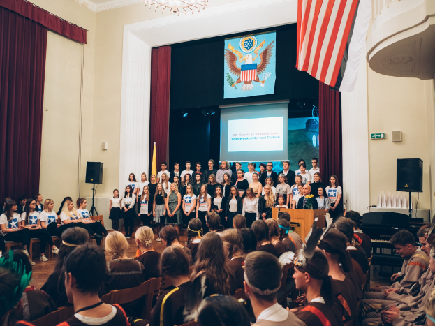 Eine Gruppe von Menschen steht an einem Podium mit einem Mikrofon, flankiert von Fahnen und Lautsprechern, die eine sitzende Menge ansprechen, während im Hintergrund ein Bildschirm, Vorhänge und ein Kronleuchter zu sehen sind.